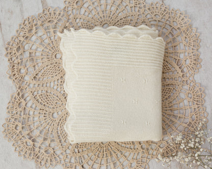 An off-white knitted baby blanket with a scalloped edge, folded and presented on a beige lace doily with small white baby's breath flowers.
