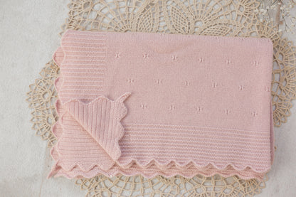 A pale pink knitted baby blanket with a scalloped edge and small embroidered star details, laid flat on a beige lace doily.